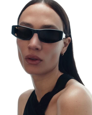 Close-up portrait of a woman wearing black rectangular sunglasses and a black halter top against a plain white studio background.