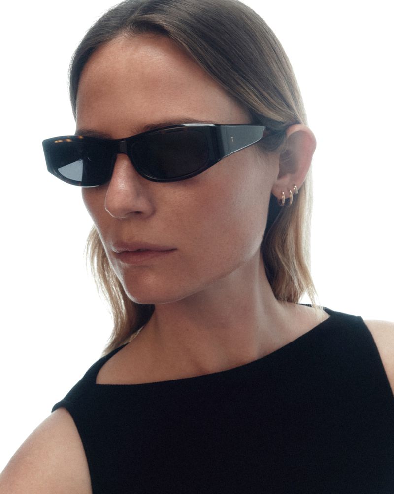 Close-up portrait of a woman wearing black angular sunglasses and gold hoop earrings against a bright white background.