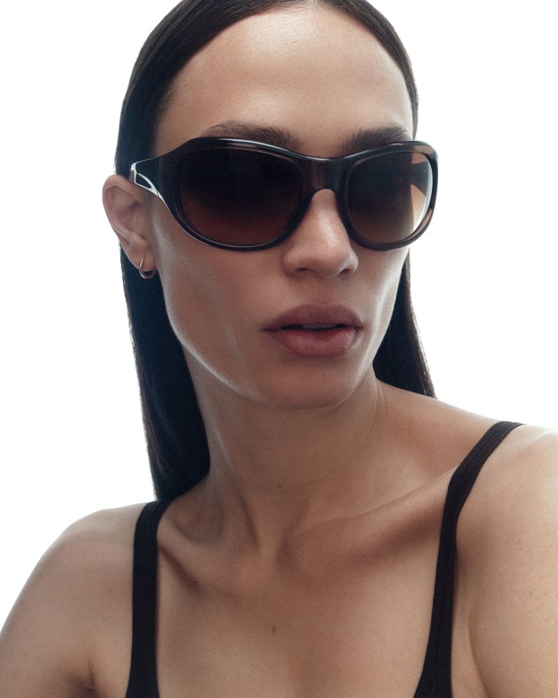 Close-up portrait of a woman wearing oversized dark sunglasses and a black tank top against a bright white background.