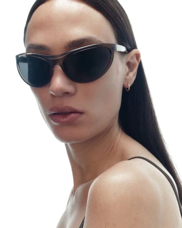 Close-up portrait of a woman wearing dark oval sunglasses with translucent frames against a seamless bright white background.