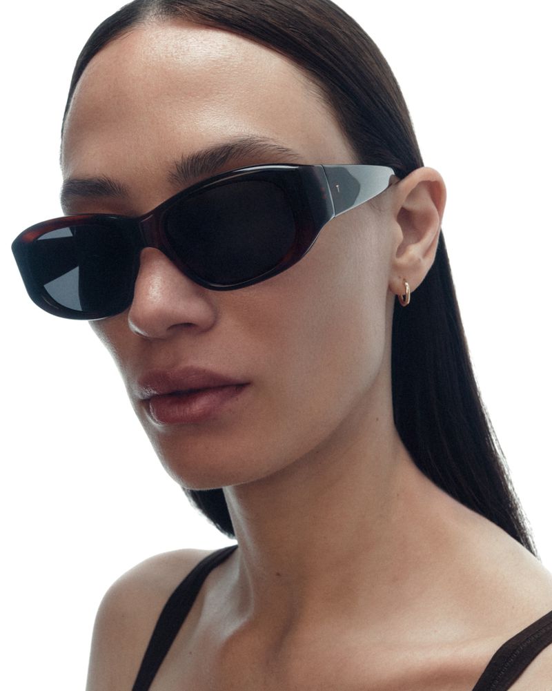 Close-up of a model wearing dark angular sunglasses and a small gold hoop earring against a plain white background.