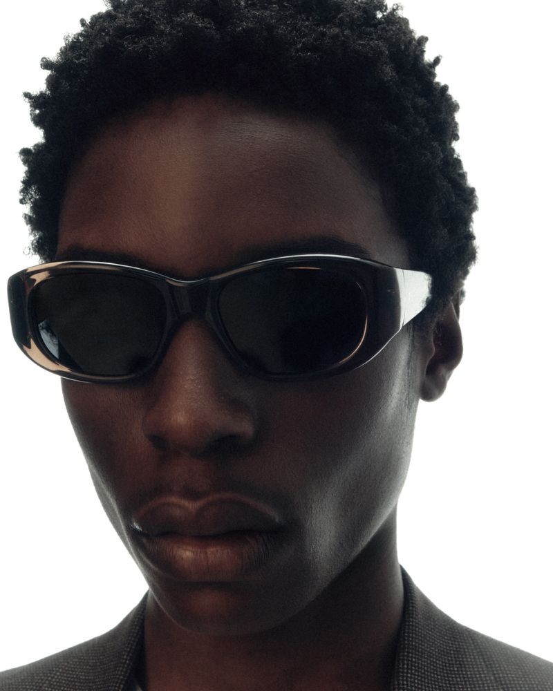 Close-up of a dark-skinned adult wearing sunglasses and a gray suit jacket against a bright white background in a studio.