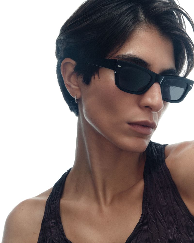 Close-up of a person with short black hair wearing black rectangular sunglasses and a dark textured top against a white background.