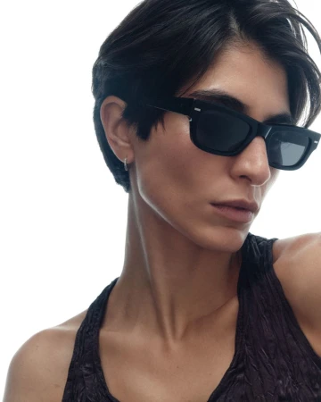 Close-up of a person with short black hair wearing black rectangular sunglasses and a dark textured top against a white background.