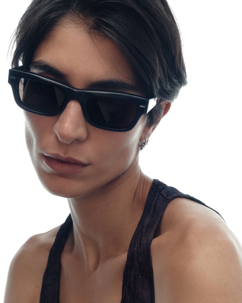 Close-up portrait of a woman with short dark hair wearing bold black rectangular sunglasses and a dark halter top against a white studio background.