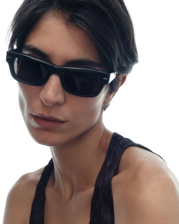 Close-up portrait of a woman with short dark hair wearing bold black rectangular sunglasses and a dark halter top against a white studio background.