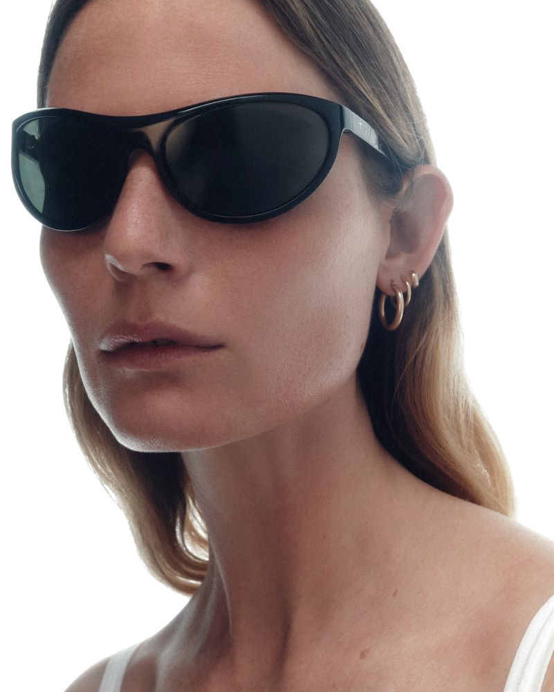Close-up portrait of a woman wearing sleek black wrap sunglasses and gold hoop earrings against a white studio background.