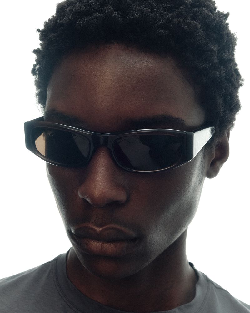 Close-up of a Black model wearing thick black rectangular sunglasses and a grey crewneck t-shirt against a white background.
