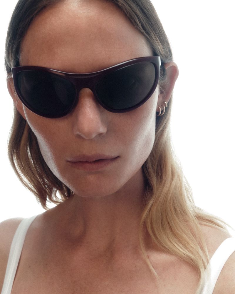 Close-up portrait of a woman wearing dark wraparound sunglasses and a white tank top against a bright white background.