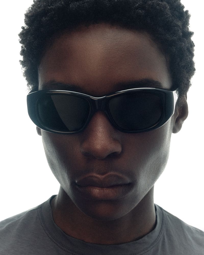 Close-up portrait of a Black model wearing thick black oval sunglasses and a grey crewneck against a white background.