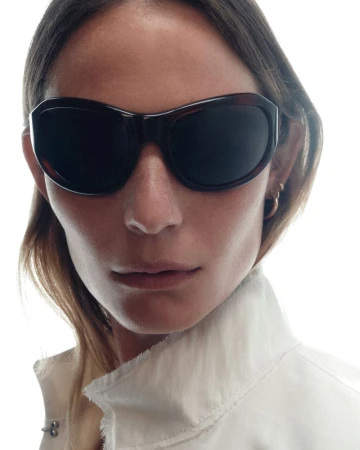 Close-up portrait of a woman wearing thick dark sunglasses and a white collared jacket against a bright white background.