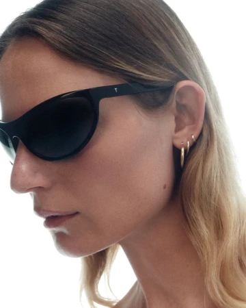 Close-up profile of a woman wearing black wrap sunglasses and gold hoop earrings against a white seamless background.