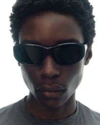 Close-up portrait of a Black model wearing thick black oval sunglasses and a grey crewneck against a white background.