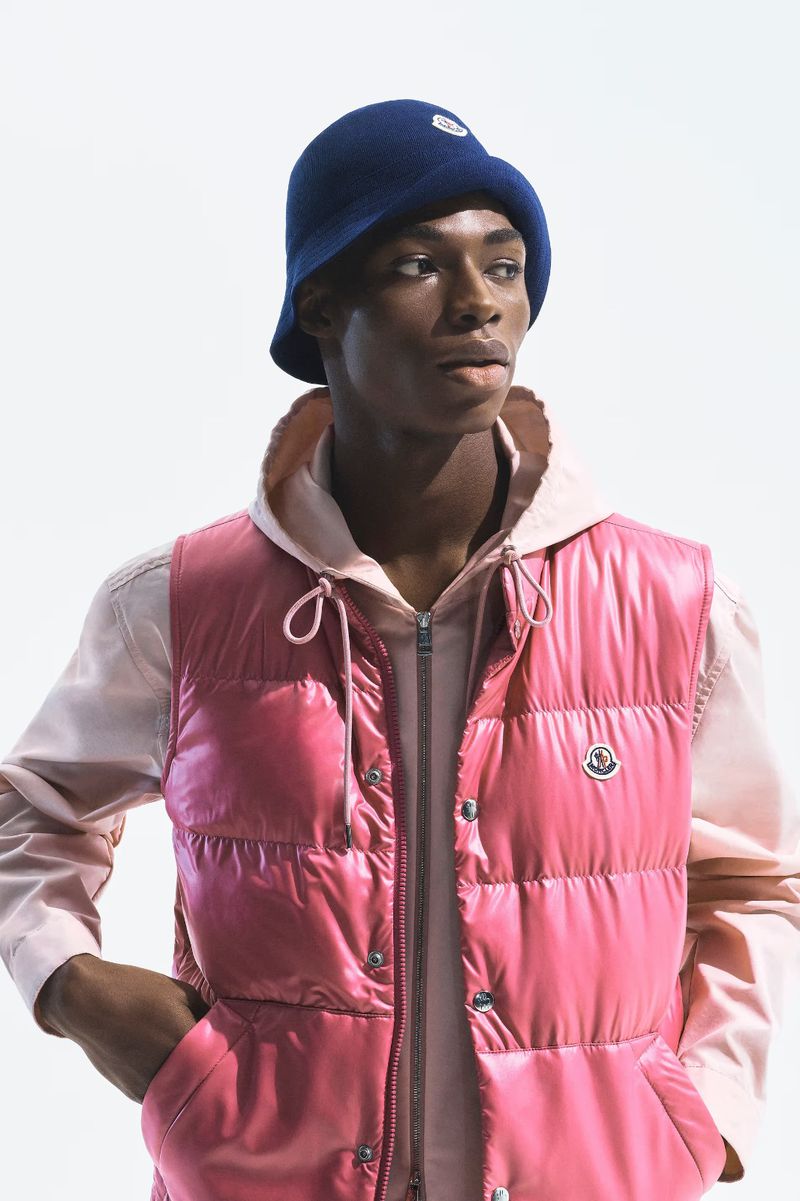 Model Zaram Obasi wearing a glossy pink Moncler puffer vest, pale pink hoodie, and navy bucket hat against a plain white studio backdrop.