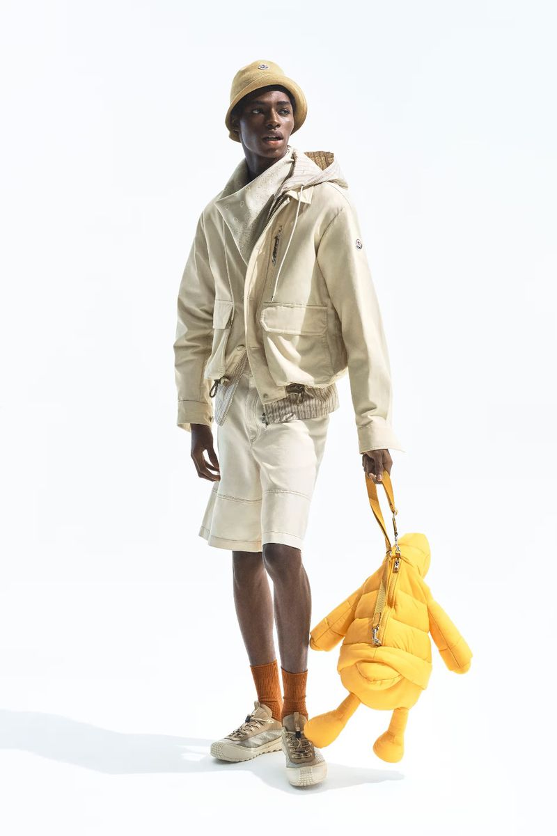 Model Zaram Obasi wearing an off-white hooded jacket, shorts, bucket hat, and holding a yellow puffy bag on a white backdrop.