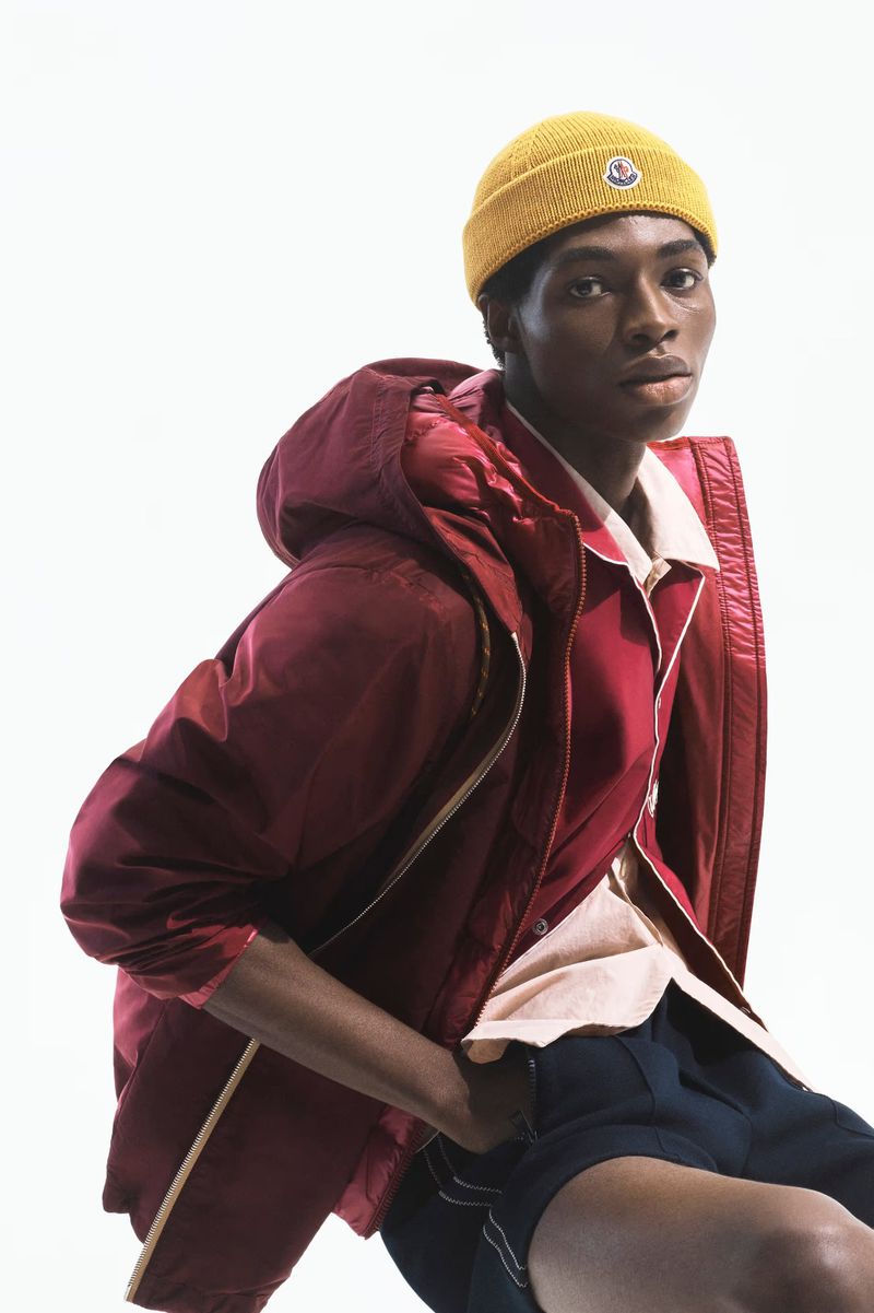 Model Zaram Obasi wearing a yellow beanie, burgundy hooded jacket, and dark shorts against a bright white seamless background.