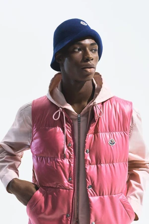 Model Zaram Obasi wearing a glossy pink Moncler puffer vest, pale pink hoodie, and navy bucket hat against a plain white studio backdrop.