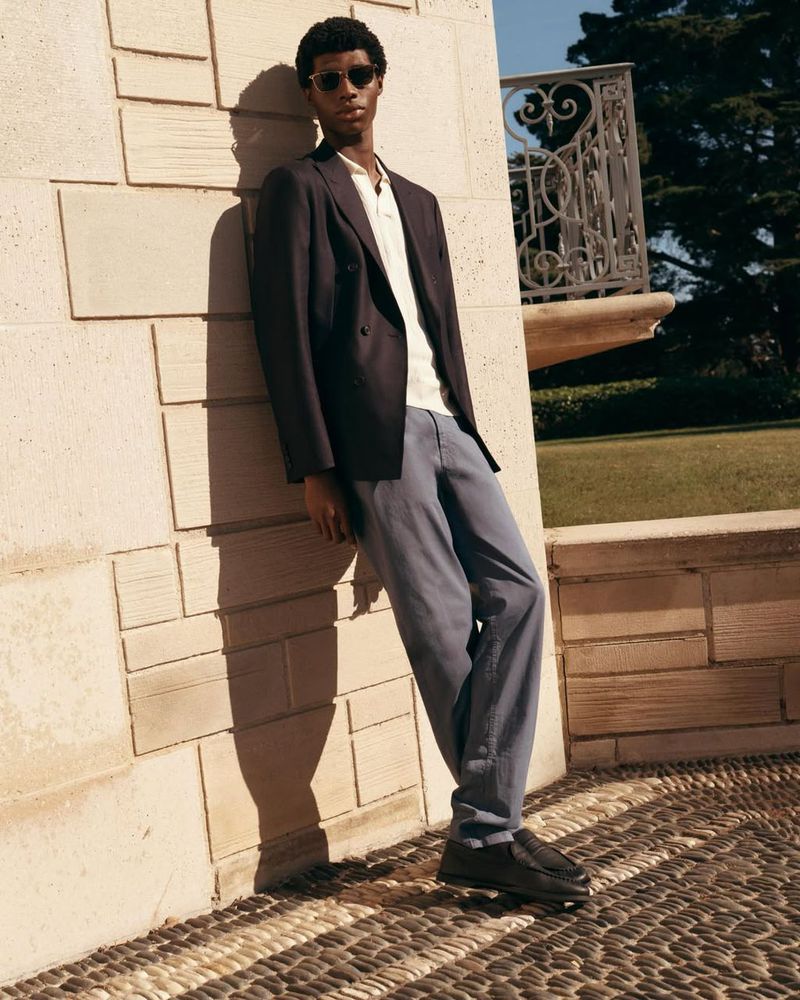 Model Zaram Obasi leans against a sunlit stone wall wearing a dark blazer, light polo shirt, grey trousers, and sunglasses.