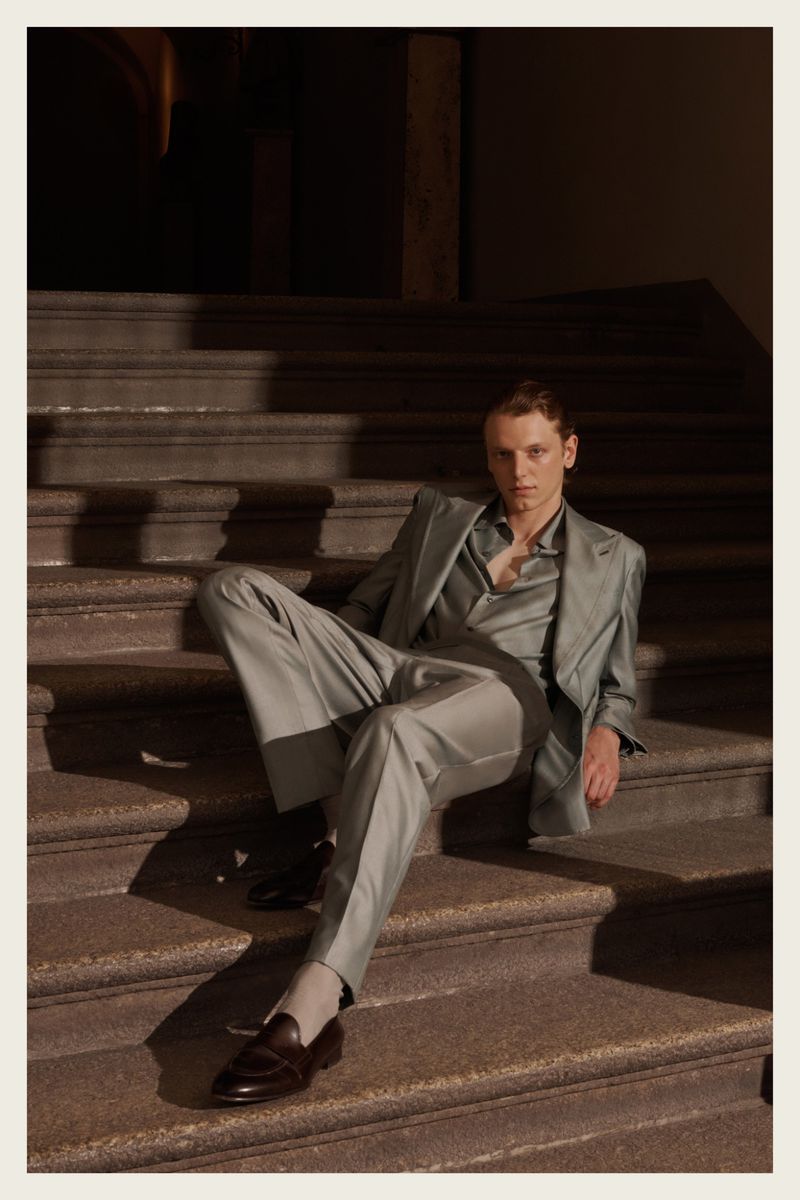A stylish man in a grey suit is sitting on stone steps in a low-light environment, showcasing fashion and architectural elements.