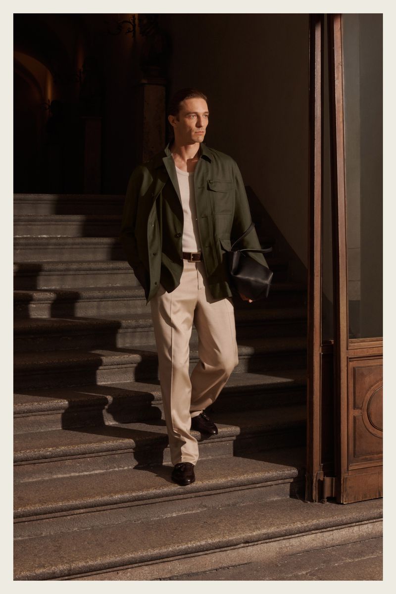 A stylish man walks down steps in an olive shirt-jacket and beige pants holding a black bag in a dim, neutral-toned, indoor environment.