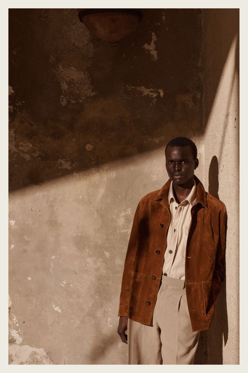 Portrait of a dark-skinned man in a brown suede jacket and cream slacks, posing against a neutral textured wall with dramatic lighting.