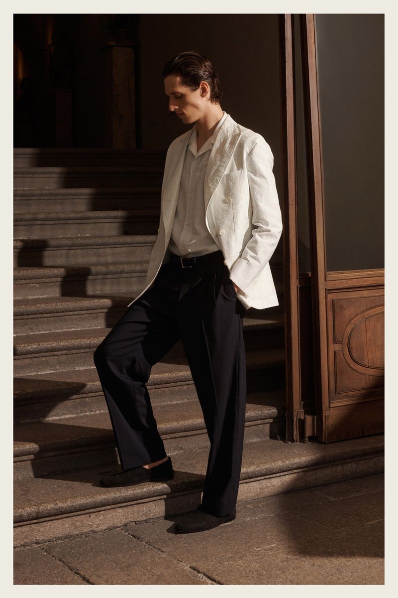 A stylish adult male stands on stone steps wearing a white jacket, collared shirt, dark trousers, and black belt in natural light.