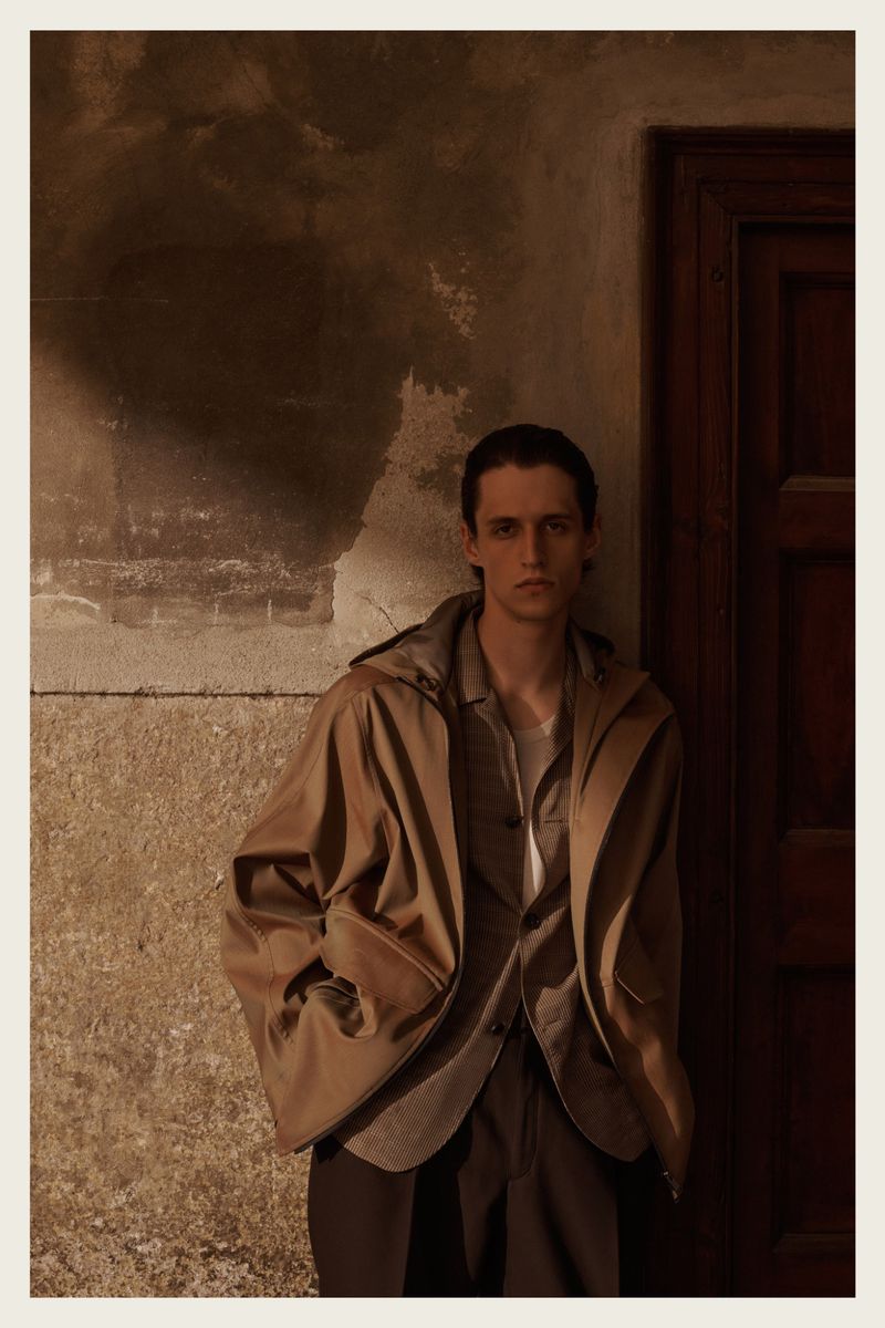 Young man in a brown hooded coat standing against an old wall near a doorway in natural light, creating a dramatic fashion portrait.