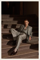 Male model in a grey tailored suit seated on stone steps outdoors in natural sunlight for Brioni Spring Summer 26.