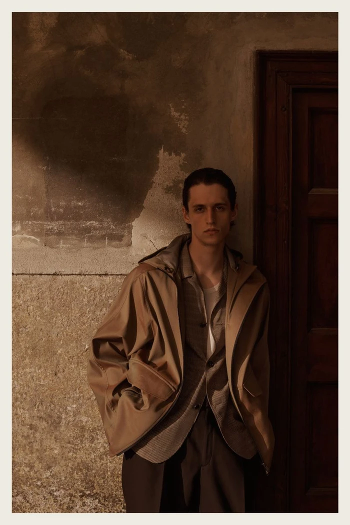 Man wearing a brown jacket standing against a textured plaster wall next to a dark wooden door.