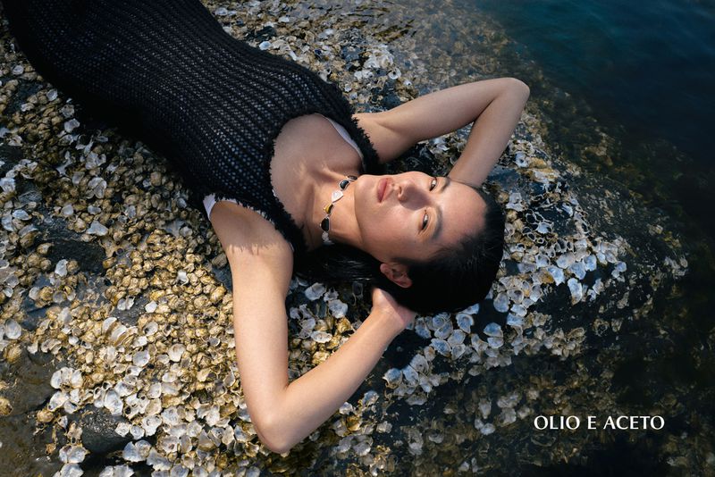 Woman in a black crochet top and necklace lying on barnacle-covered rocks by the water for Olio E Aceto.