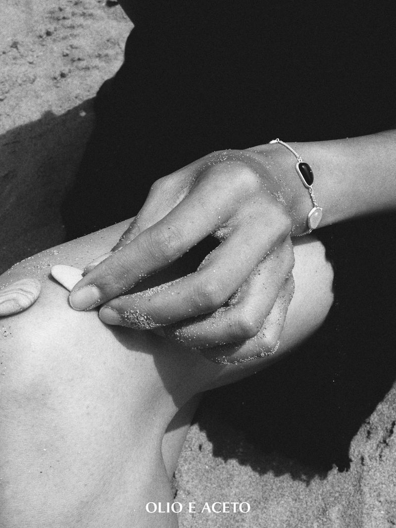 Close-up black and white shot of a sand-covered hand wearing a delicate stone bracelet placing pebbles on a knee.