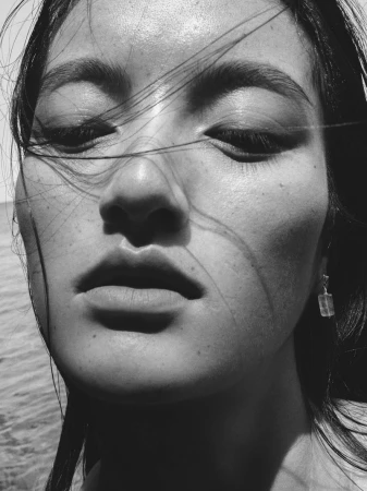 Close-up black and white portrait of an East Asian woman with loose hair blowing across her face outdoors.