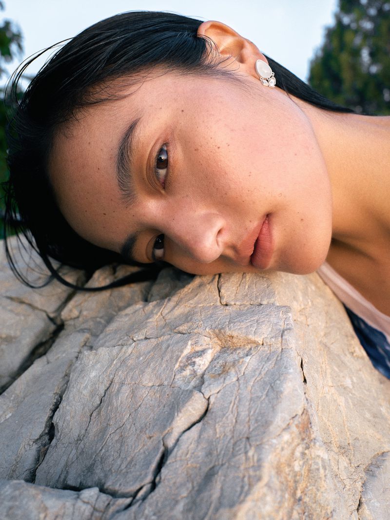 Close-up of an East Asian woman resting her head against a textured rock surface wearing a white earring.