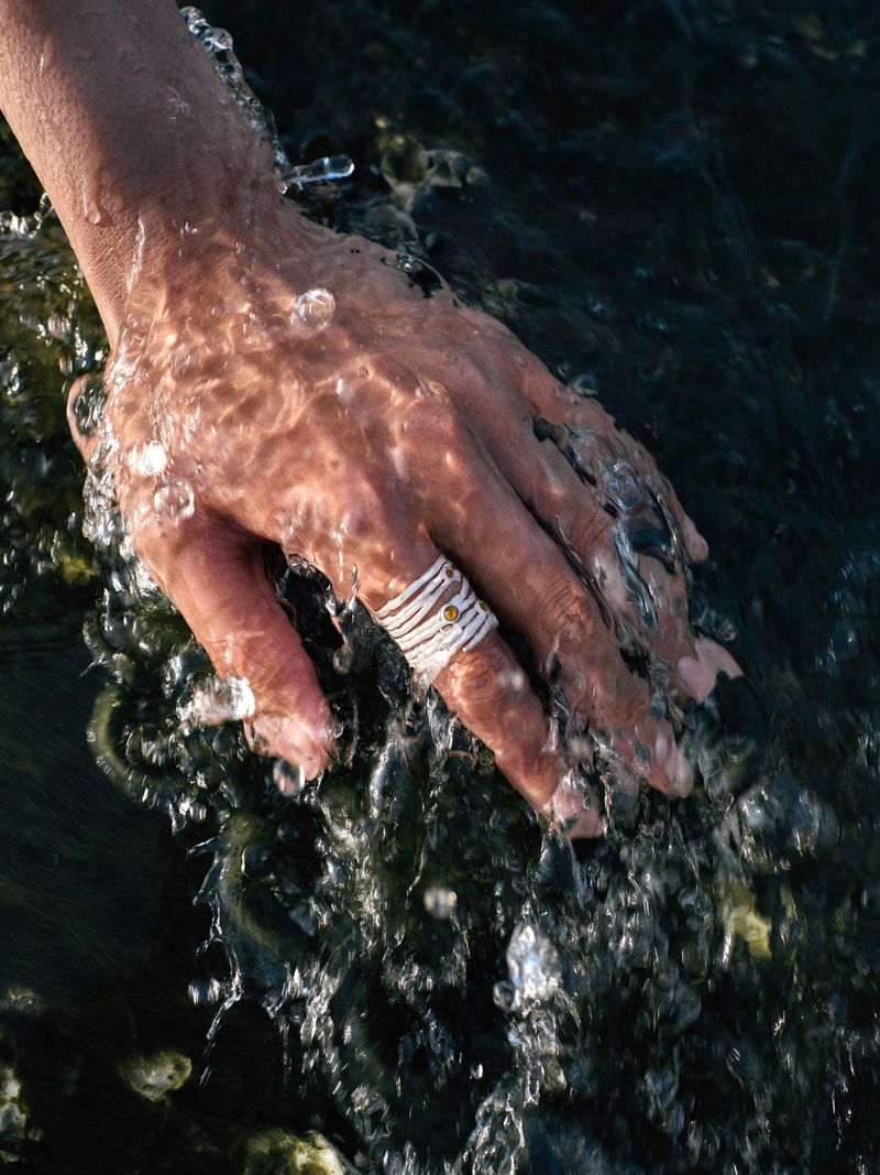 Close-up of a hand reaching into splashing dark water wearing a white multi-band wrap ring with a small amber stone.