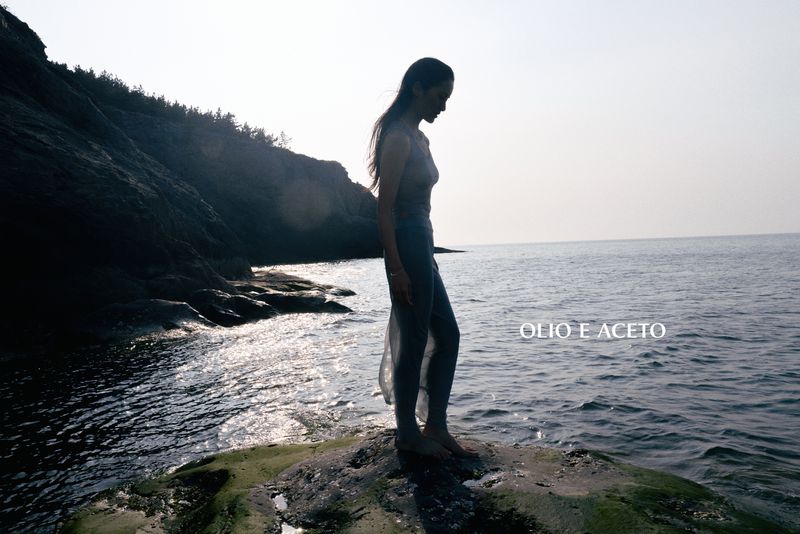 A woman stands barefoot on a moss-covered rock at the rocky ocean shoreline against bright backlit sky.