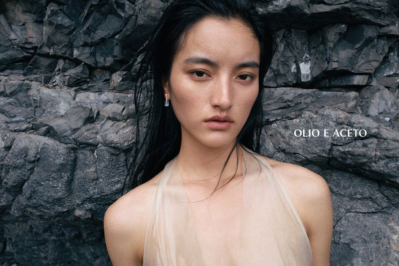 East Asian model with wet black hair wearing a sheer beige halter top against a dark, rugged rocky cliff face.