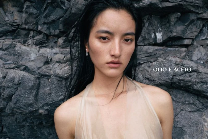 East Asian model with wet black hair wearing a sheer beige halter top against a dark, rugged rocky cliff face.