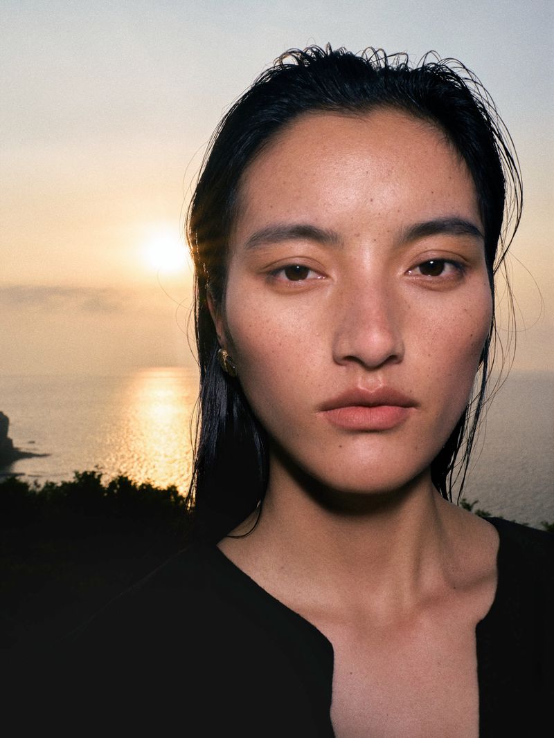 Close-up portrait of an East Asian woman with slicked-back wet hair in front of an ocean sunset.