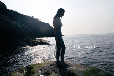 A woman stands barefoot on a moss-covered rock at the rocky ocean shoreline against bright backlit sky.