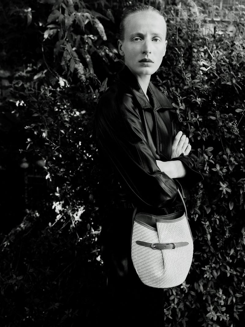 Monochrome portrait of a model in a dark jacket holding a woven shoulder bag in front of foliage.