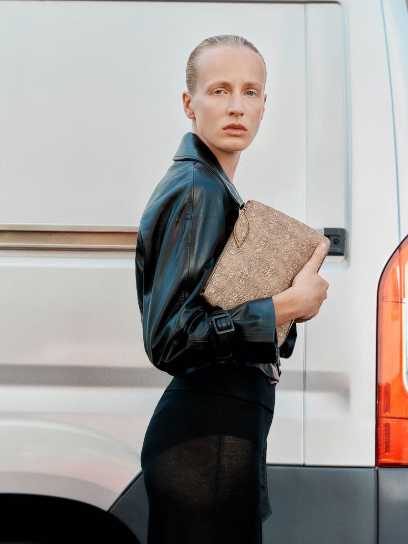 Model Anine Van Velzen in a black leather jacket holding a beige patterned clutch against a white vehicle.