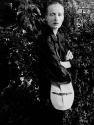 Monochrome portrait of a model in a dark jacket holding a woven shoulder bag in front of foliage.