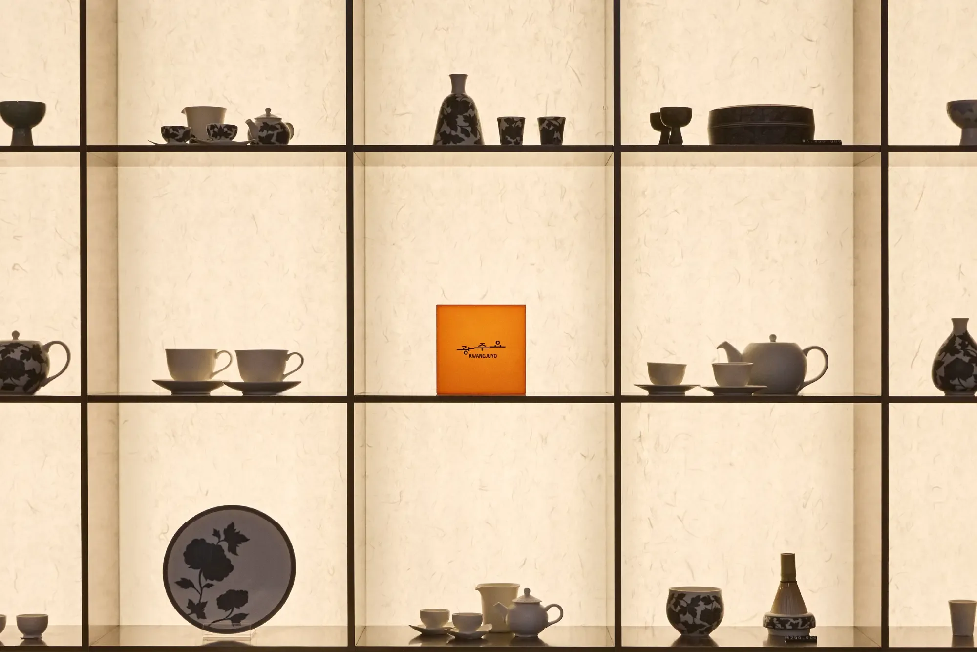 Ceramic teapots, cups, and vessels displayed on backlit grid shelving with an orange Kwangjuyo logo box in the center compartment.