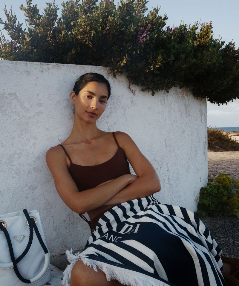 A brunette woman in a brown top poses with arms crossed while seated next to a white Prada tote bag in an outdoor environment.