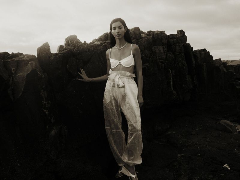 Woman poses in sheer pants and a bra top against a rocky shoreline with a cloudy sky, creating a neutral, textured fashion image.