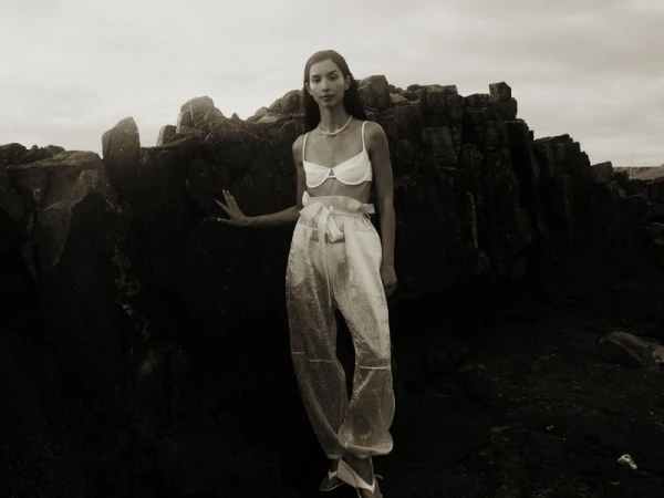 Woman poses in sheer pants and a bra top against a rocky shoreline with a cloudy sky, creating a neutral, textured fashion image.