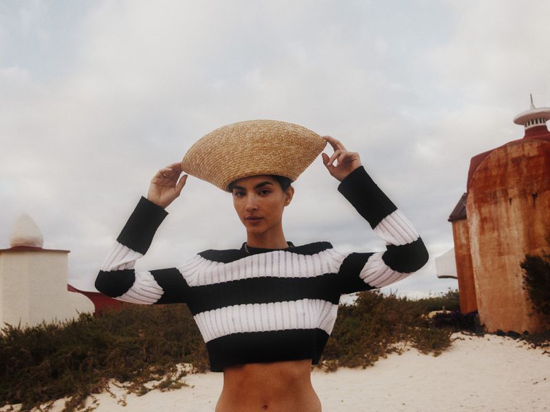 A woman poses outdoors on a sandy beach wearing a striped shirt and straw hat, with red and white buildings in the background.