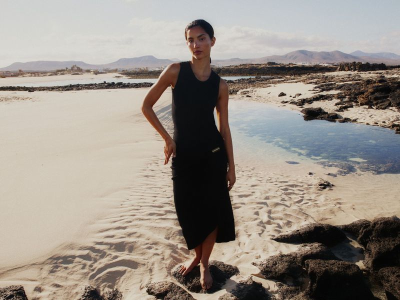 A woman in a black dress poses barefoot on a sandy beach near rocky tide pools with a mountainous horizon under a bright sky.