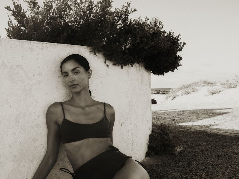 Monochrome shot of a woman in a bikini leaning against a wall with vegetation, with a coastal landscape visible in the background.
