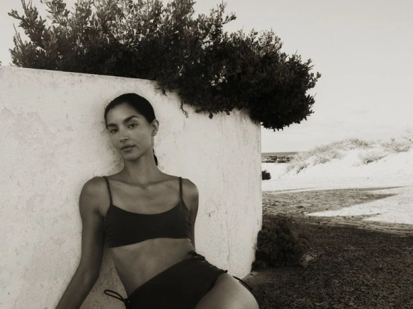 Monochrome shot of a woman in a bikini leaning against a wall with vegetation, with a coastal landscape visible in the background.